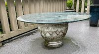 Full side view of round glass top patio table with decorative base in outdoor setting.