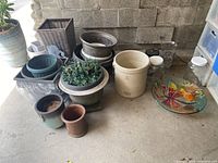 Overview of all items including multiple plant pots of ceramic and plastic, large crock, glass dish with floral design, glass bottles, faux wicker planter, and metal garden figure