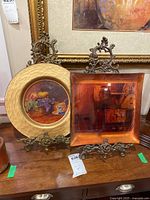 Two plates on ornate bronze metal stands, one round with painted fruit and cup design with hammered gold rim, the other square with glossy brown-orange finish.