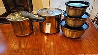 Photo showing the stainless steel pressure cooker with black handle, large stock pot, and the set of four nested stainless steel bowls with black lids, all on a wooden table.
