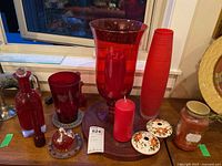 Photo showing red glass items on a wooden surface near a window, including large vase, candle holder, candle, decorative lids, and a candle holder base plate.