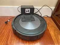 iRobot Roomba j7 robot vacuum resting on the black docking/charging station on hardwood floor, visible power cord attached to dock.