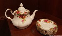 Teapot and stack of seven saucers arranged on a wooden table showing floral patterns and gold trim on the china.