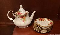 Royal Albert Old Country Roses teapot displayed alongside a stack of nine matching saucers with floral design and gold trim.