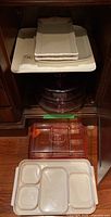 Stack of white Life Home ceramic serving plates placed on top of white plastic trays, with clear plastic trays and lids below in wooden cabinet