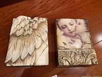 Two Sid Dickens wall mounts laid side by side on a wood floor. Left plaque is Wing with sculpted wings. Right plaque is The Kiss with painted image.