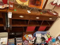 Wood sideboard with three drawers and six open cubby compartments, dark wood finish with scratches and scuffs, shown in room with various items around it.