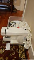 Front view of white Singer sewing machine Model 132Q with red Singer logo, foot pedal and white fabric inside a plastic bin behind it.