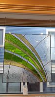 Full view of square stained glass panel with green, blue, clear, and amber strips, hung and photographed against a window showing a residential building outside.