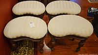 Four wooden stools with kidney-shaped upholstered seats shown from top angle, light-colored fabric with marks visible.