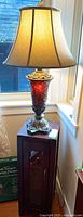 Full view of the table lamp on top of a dark wood cabinet next to a window. Shows lamp shade and mosaic glass base.
