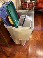 Plastic storage totes stacked inside a large clear plastic bin on a hardwood floor with various colored lids on top (green, yellow, maroon, blue, purple).