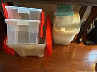 Two groups of plastic storage totes on a wooden floor under a table. Left group has clear containers with red lids stacked together, right group has translucent white storage totes with clear lids.