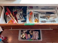 Top view of a drawer with divided sections containing various kitchen utensils including scissors, peelers, whisks, measuring spoons, pizza cutter, garlic press, and scrub brushes.