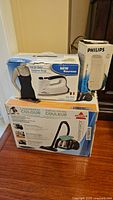 Three boxed items on hardwood floor: Philips steamer, Euro Steam iron, Bissell vacuum