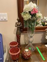 Tall glass vase with faux pink and white flowers, two red Venetian ceramic plant pots, round red and gold ceramic tealight holder, and decorative figurine on a glass surface in front of a mirror.