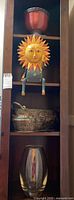 Shelf with a reddish-brown ceramic planter on top, wooden sun figure in middle, heavy metal plant pot below, and clear glass vase at bottom