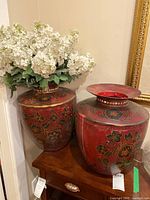 Two red decorative metal vases with floral designs placed on wooden surface, the left vase holding white faux hydrangea flowers. The vases show visible scratches and scuffs.