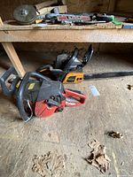 Side view of two chainsaws on wooden floor under a bench. Poulan Pro chainsaw is complete with chain and bar. Jonsered chainsaw missing the bar and chain.