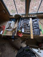 Top-down image of cordless drill with battery and hand tools laid out on wooden surface near window, showing variety of wrenches, pliers, chisels, drill bits, and a level.