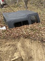 Black truck cap partially buried in leaves and dirt, showing one side with tinted sliding windows.