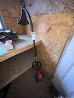 Full view of a red Homelite gas-powered weed eater with curved shaft leaning against a wooden wall, showing the handle, protective guard, and engine area.