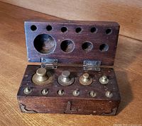 Wooden chest open showing ten brass weights placed in fitted holes inside lid and base.