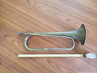Full side profile of antique brass bugle next to measuring tape for scale on wooden floor.