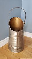 Front side view of the metal antique coal scuttle showing the broad top opening and the handle raised upwards, placed on wooden floor against grey wall.