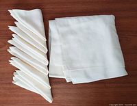 Folded 46 inch white antique tablecloth alongside six neatly folded matching napkins on wooden surface.