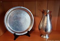Photo shows the set of a large hammered textured Indonesian pewter jug and round serving tray on wooden shelf.