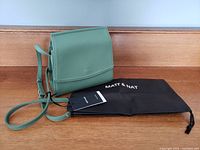 Green Matt and Nat purse sitting atop its black protective bag with attached tag visible, showing crossbody strap and front flap.