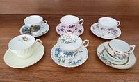 Photo showing six vintage bone china teacups and saucers from multiple prestigious brands arranged side by side on wooden surface.