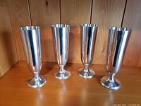 Four tall cocktail glasses made from polished Indonesian pewter standing in a row on a wooden shelf.