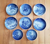 Eight Royal Copenhagen Christmas plates arranged on wooden surface showing detailed traditional blue and white winter holiday scenes.