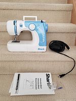 Sewing machine with attached black foot pedal cord and instruction manual with spare presser feet laid out on carpeted stairs.
