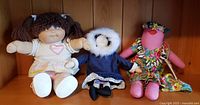 Three dolls on wooden shelf: vintage Cabbage Patch signed by Xavier Roberts, Inuit rag doll with fur-lined hood, Pacific Island rag doll with floral dress.