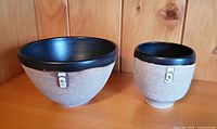 Two pottery bowls on wood surface against wood panel background, showing size and appearance of black interior and taupe textured exterior with decorative element