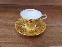 Front angled view of vintage gold yellow Aynsley teacup and saucer set showing ruffled edges and detailed gold patterns.