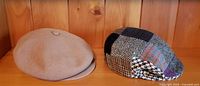 Two caps displayed on a wooden shelf, one camel color Kangol and one multi-pattern Irish wool patchwork.