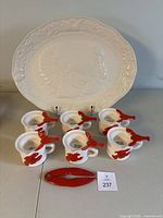 Six white ceramic lobster butter melting pots arranged in one row in front of a large off-white ceramic turkey platter with raised embossed decoration. The lobster dishes have a red lobster design on the side and a top section for butter.