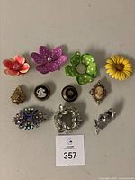 Top-down photo showing 11 assorted pins including flower brooches in various colors, cameo and vintage style brooches, and stone cluster pieces.