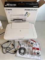 Canon Pixma MG2522 printer, original box, power cable, sealed black and color ink cartridges, setup CD, and instruction manual arranged on table.