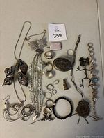 Photo showing assorted costume jewellery laid on a flat surface including necklaces, bracelets, rings, pendants, and earrings. Variety of shapes and sizes visible including floral design necklace and chunky link bracelet.