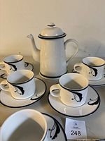 Photo showing the full set of 11 white cups with blue curved floral designs and matching saucers arranged around the white coffee pot with blue trim.