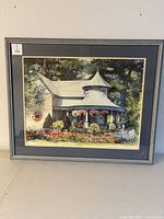 Full view of the framed watercolor painting showing a cottage surrounded by a garden with flowers. The frame is grey wood and the matting is blue with some stains or marks.