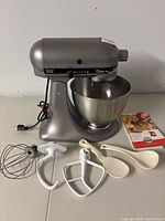 Front view of mixer on table with bowl, attachments, spatulas and booklet arranged in front