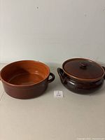 Two terra cotta cooking pots side by side, one round brown glazed pot with lid and two handles, one orange unglazed pot with two handles
