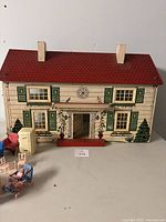 Front view of vintage wooden doll house with red tin roof, two chimneys, painted green shutters, floral decorations, and assortment of miniature furniture pieces in front.