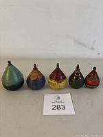 Front view of 5 vintage wooden spin tops lined up, showing various colors and metal tips, with auction number card in front